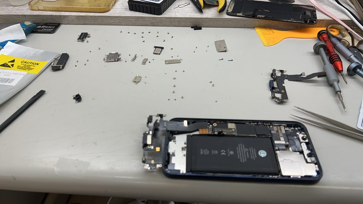 iPhone 12 拆機更換尾插排線，桌上可見拆下的舊排線與各內部零件
